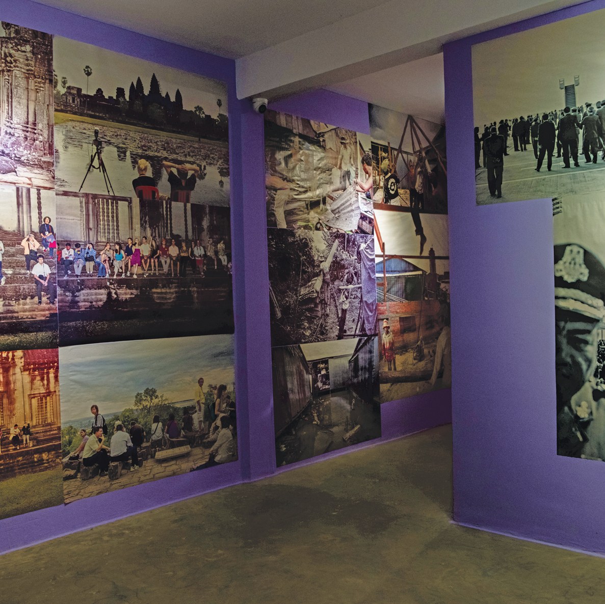 A gallery with purple walls displays large photographic prints depicting landscapes, people, historical sites, and black-and-white imagery of organized gatherings.