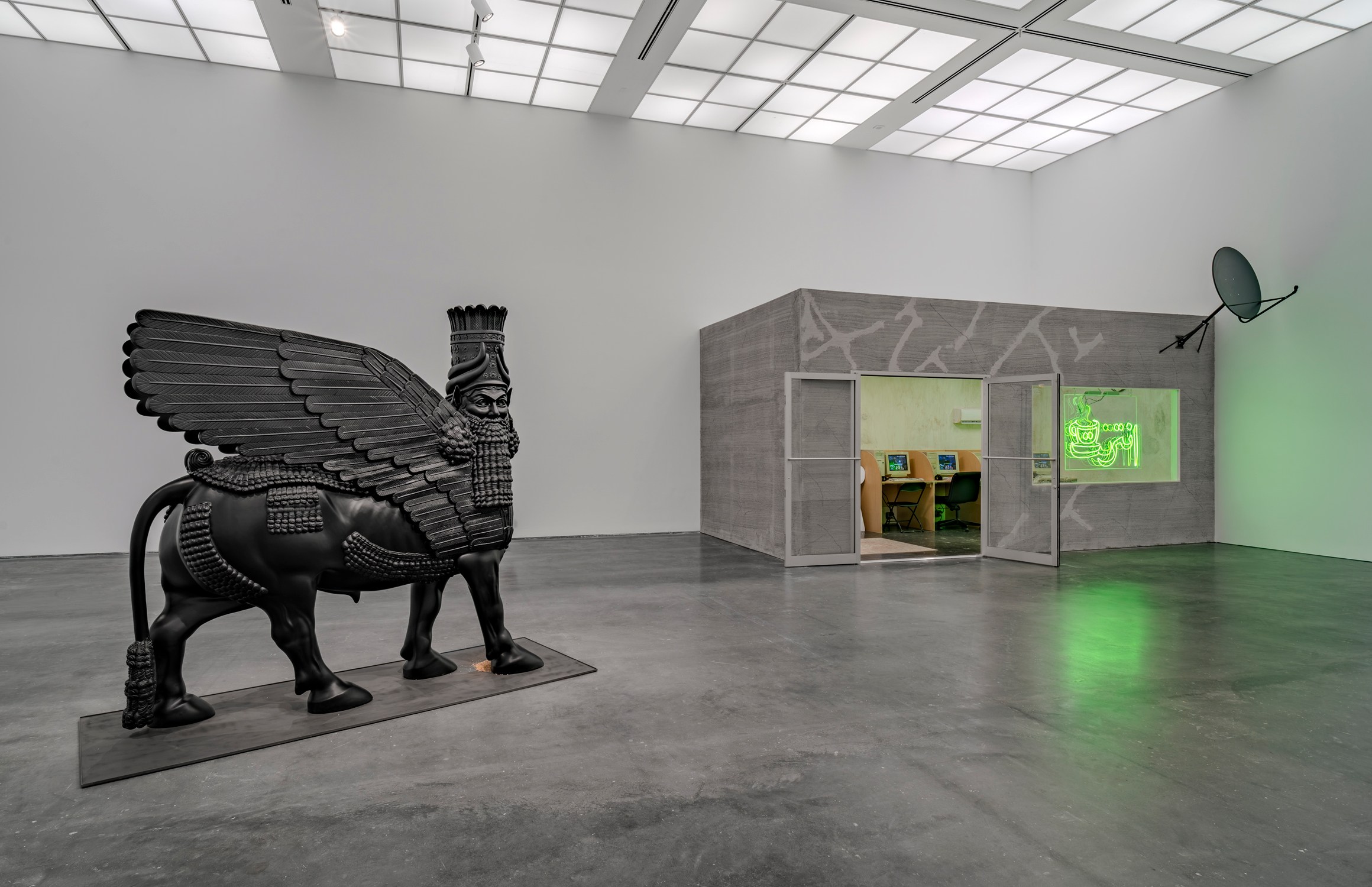 A black-winged bull sculpture stands on the left; a gray structure with doors, neon green signage, and workstations inside sits to the right. View of “Wafaa Bilal: Indulge Me,” 2025, Museum of Contemporary Art Chicago. From left: Lamassu (In a Grain of Wheat), 2025; In a Grain of Wheat: Cultivating Hybrid Futures in Ancient Seed DNA, 2021; Virtual Jihadi, 2008.