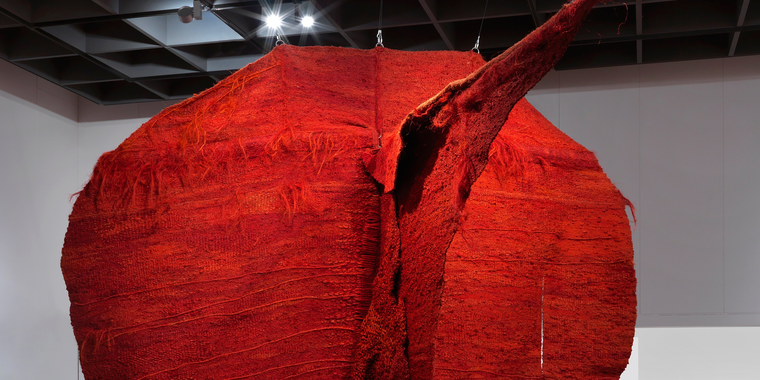 A large, suspended red textile sculpture resembling a circular form with textured surfaces and an extended tapered point, displayed in a gallery space.